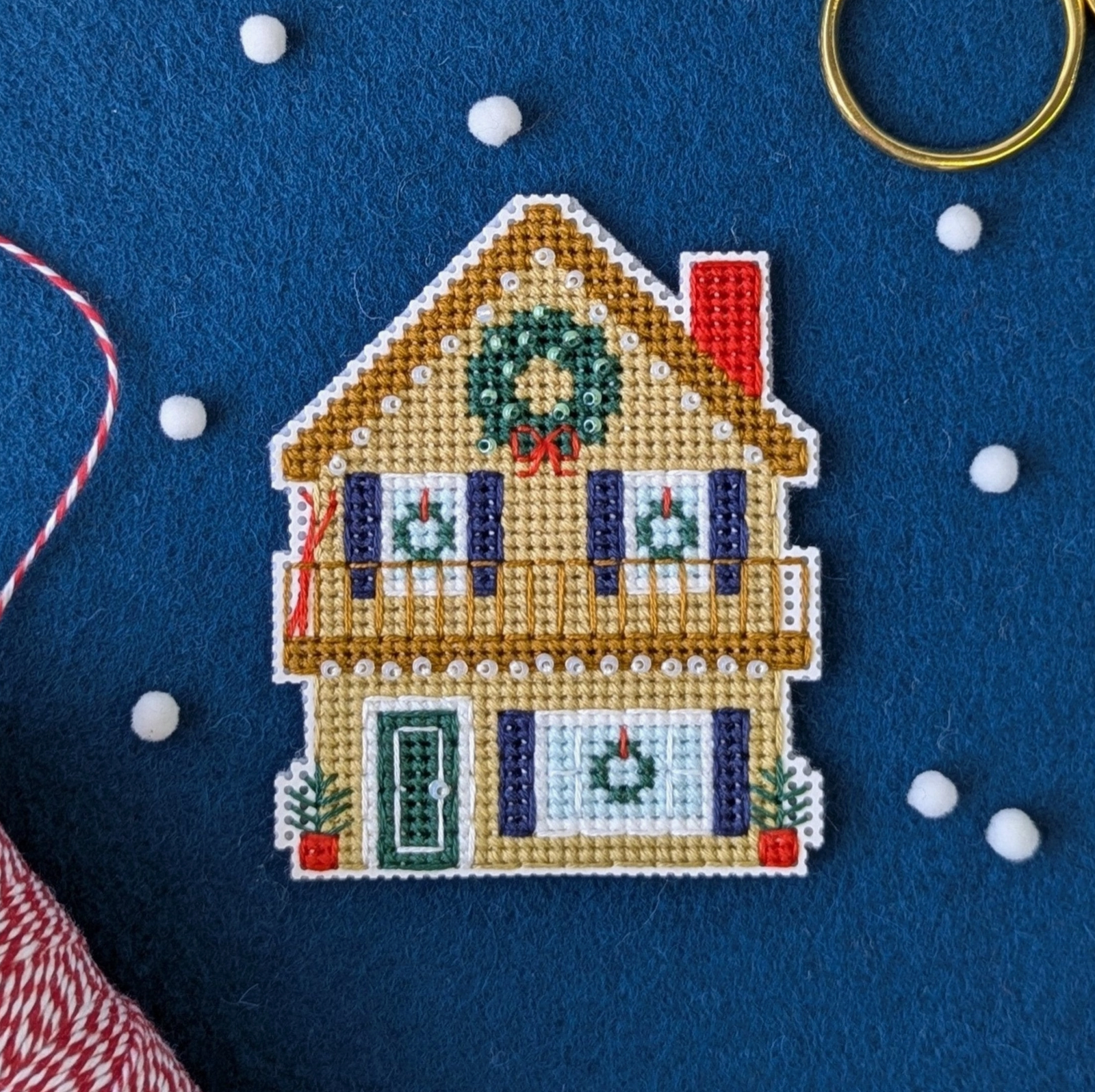 A cross stitch ornament of a holiday home chalet is sitting atop a piece of royal blue felt with some white beads scattered across the table.