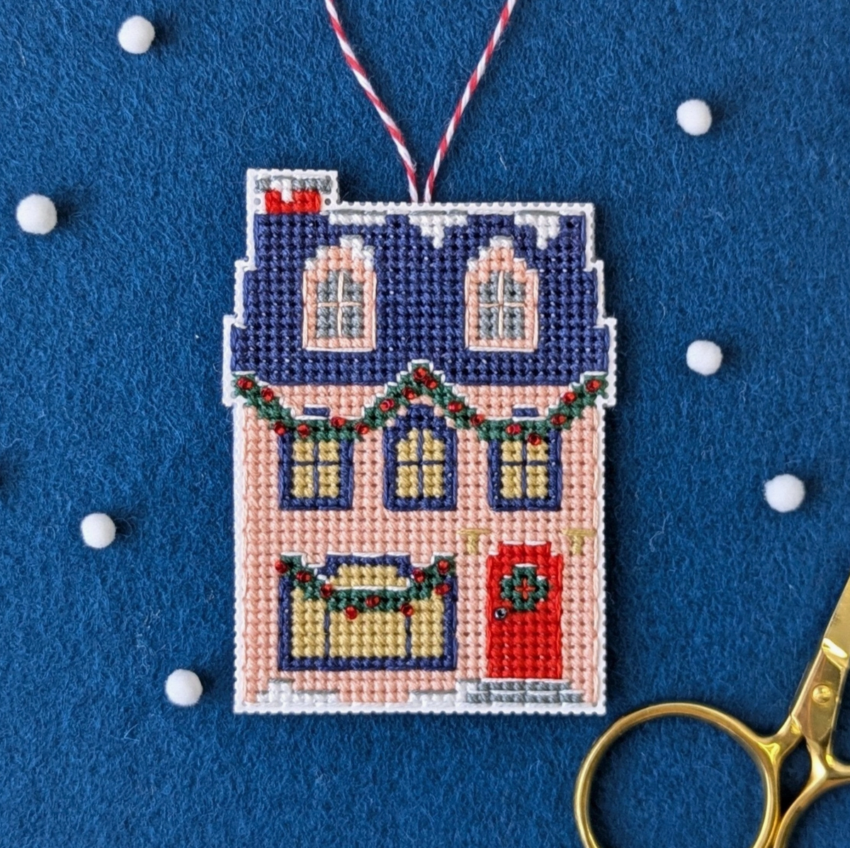 A cross stitch ornament of a holiday home cottage is sitting atop a piece of royal blue felt with some white beads scattered across the table.