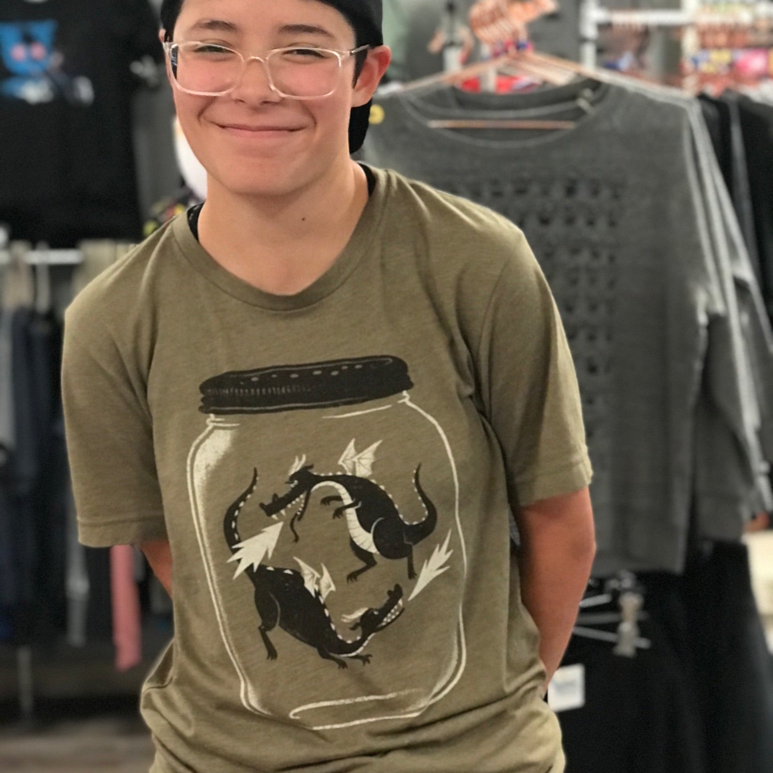 A person wearing a backwards baseball cap is smiling and squinting behind clear framed glasses. They are wearing an olive green tshirt with dragonflies on it. The dragonflies are in a jar - but they look like tiny little dragons.