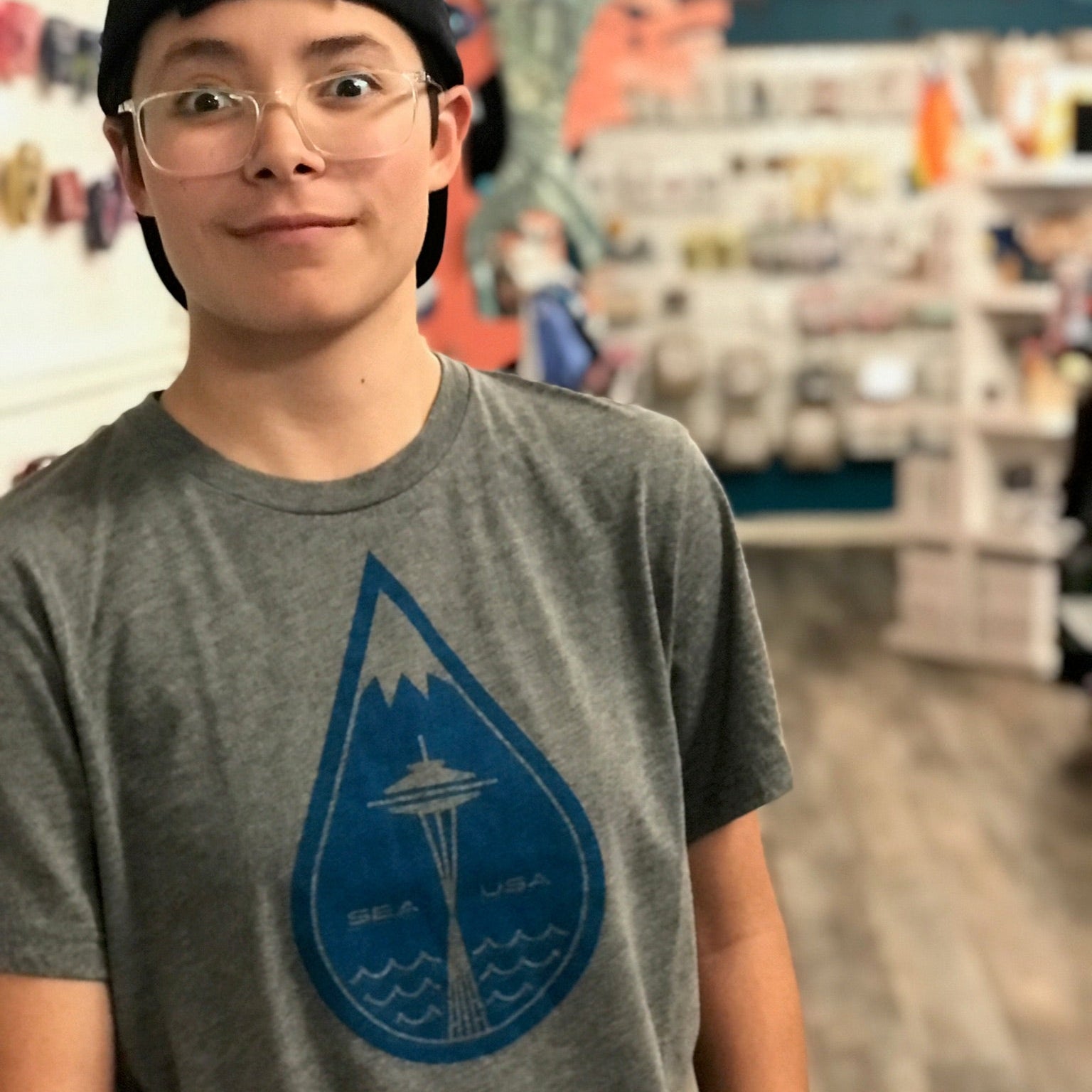 A person wearing a shirt with a big raindrop in the middle. The top of the raindrop looks like a mountain cap and there is a Space Needle in the middle. 