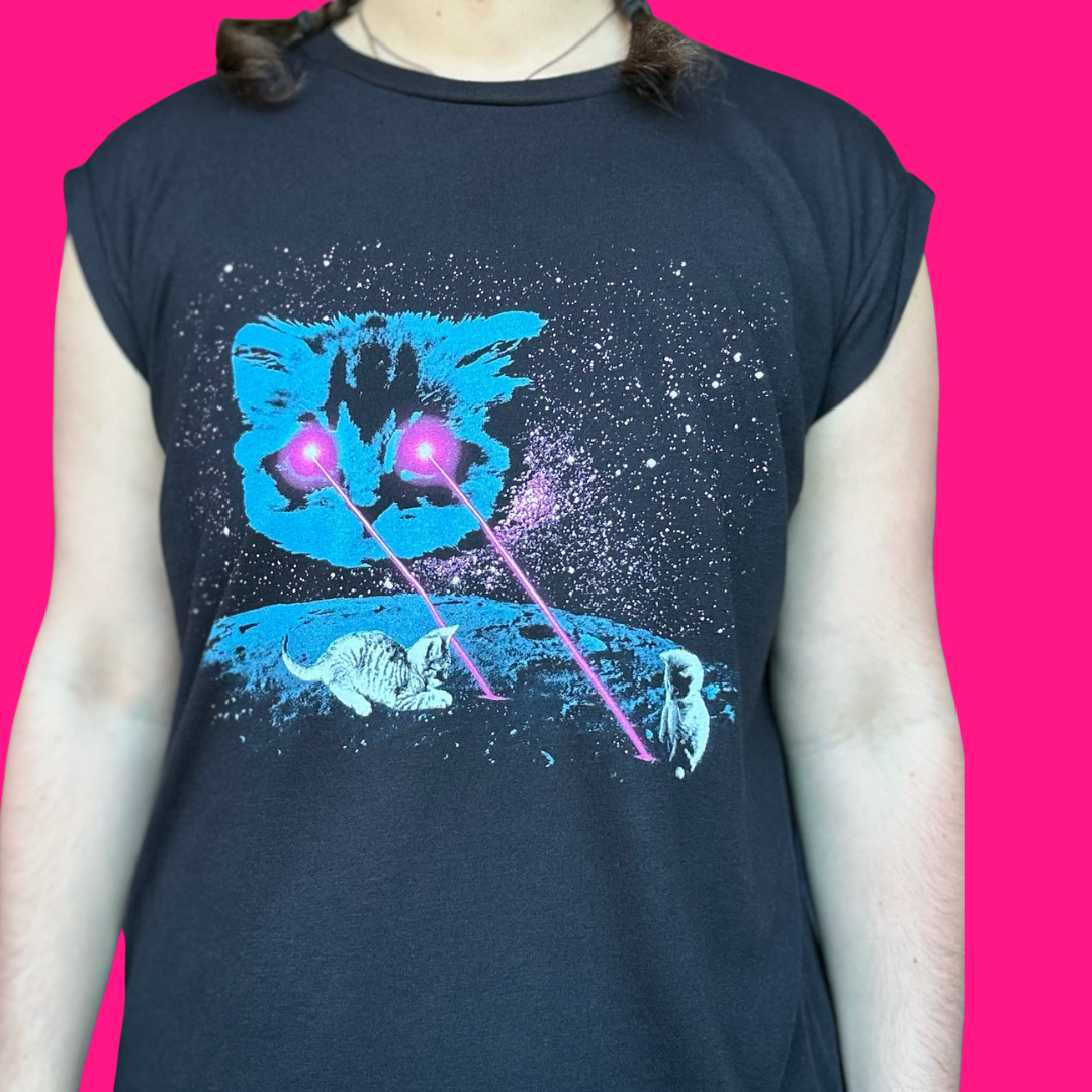 A person wearing a dark navy sleeveless t-shirt with a graphic print of a blue cat's face, white cats, and pink laser beams on a starry black background, set against a solid bright pink backdrop.