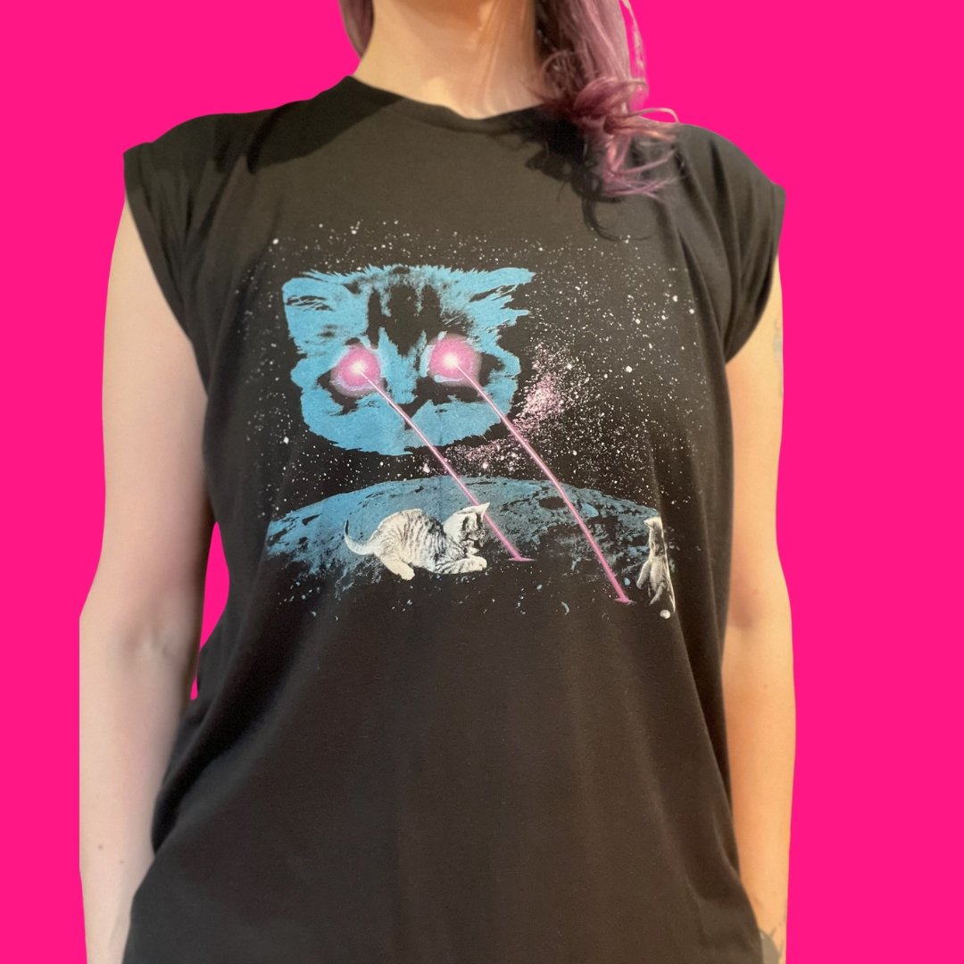 A person wearing a dark navy sleeveless t-shirt with a graphic print of a blue cat's face, white cats, and pink laser beams on a starry black background, set against a solid bright pink backdrop.