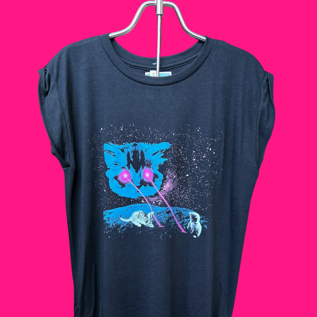 A person wearing a dark navy sleeveless t-shirt with a graphic print of a blue cat's face, white cats, and pink laser beams on a starry black background, set against a solid bright pink backdrop.