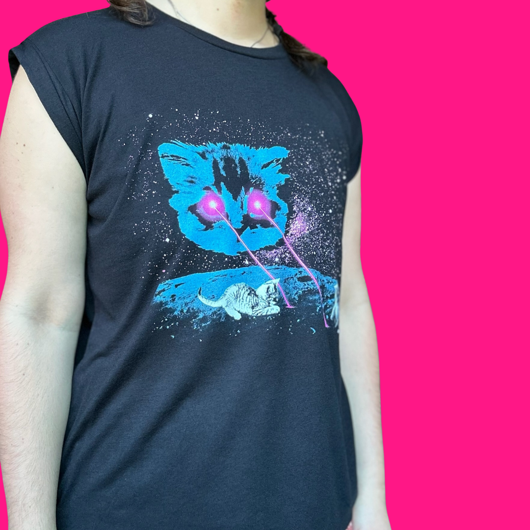 A person wearing a dark navy sleeveless t-shirt with a graphic print of a blue cat's face, white cats, and pink laser beams on a starry black background, set against a solid bright pink backdrop.