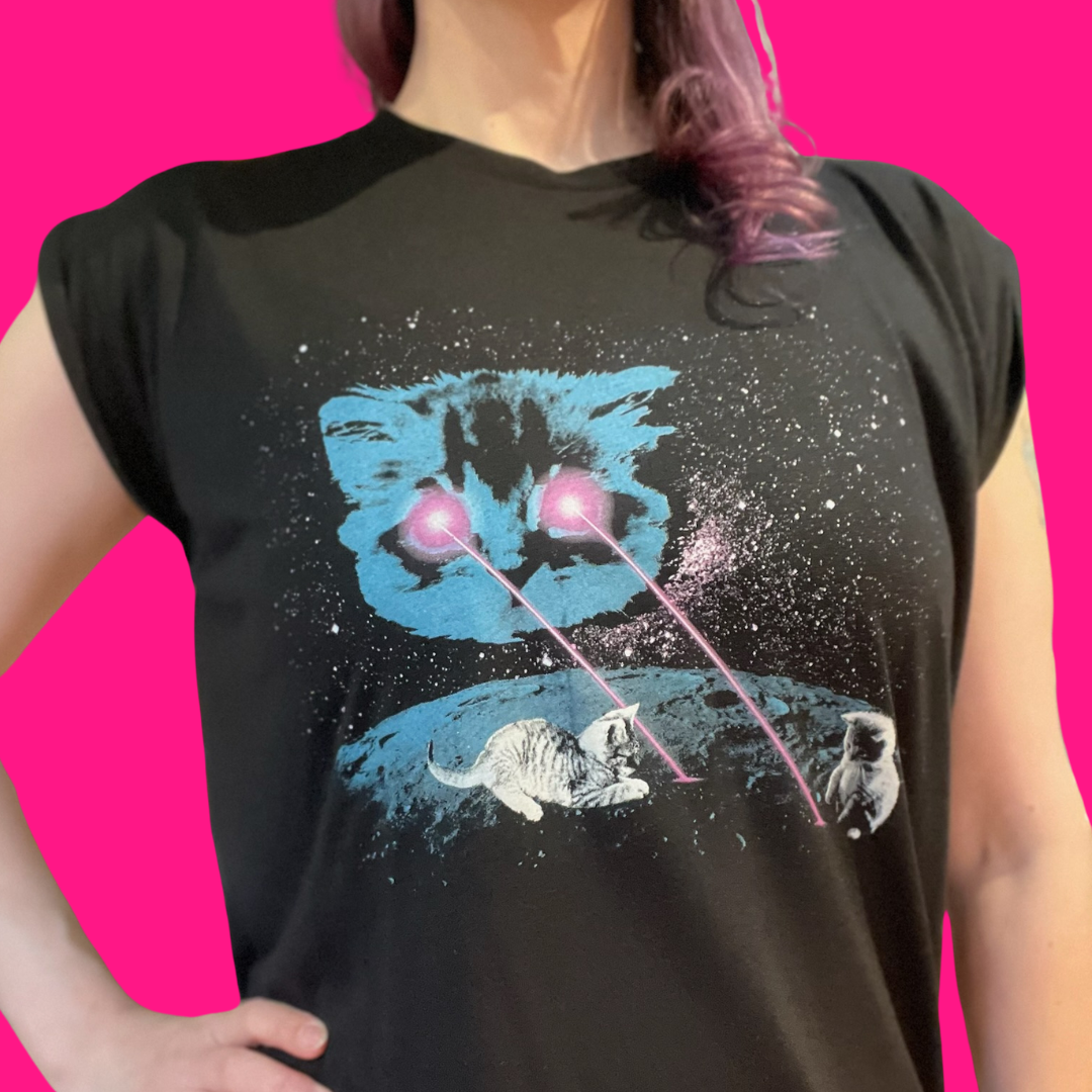 A person wearing a dark navy sleeveless t-shirt with a graphic print of a blue cat's face, white cats, and pink laser beams on a starry black background, set against a solid bright pink backdrop.
