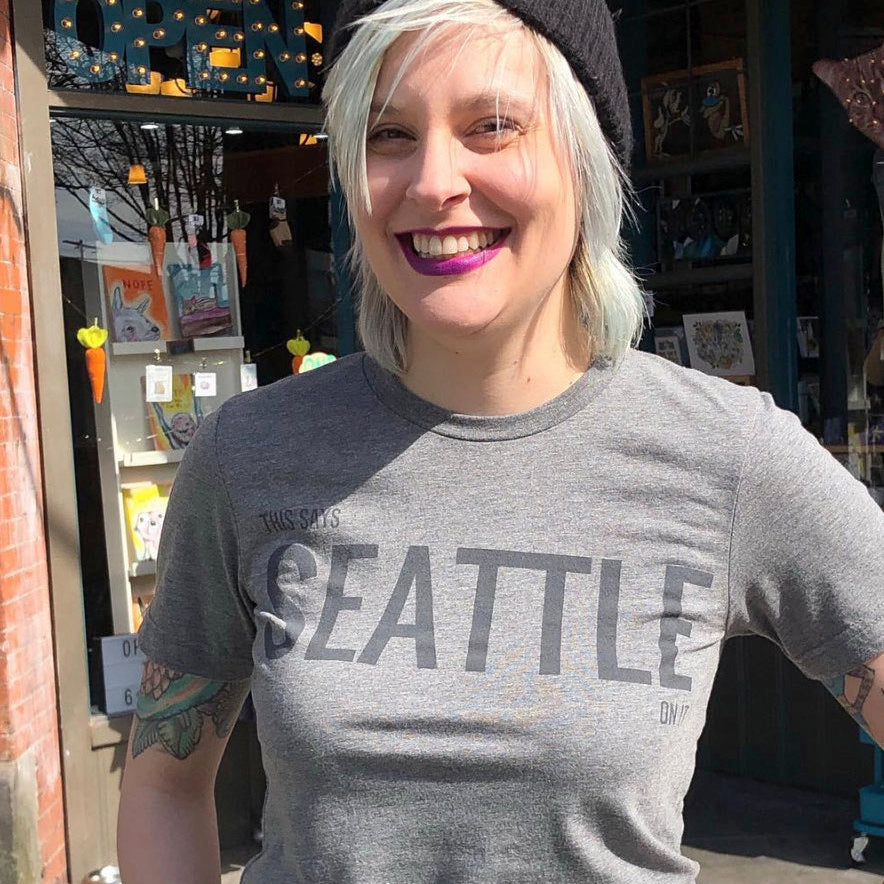 A smiling person stands outside in the sunshine. They are wearing a heather gray t-shirt that says "This Says Seattle On It."