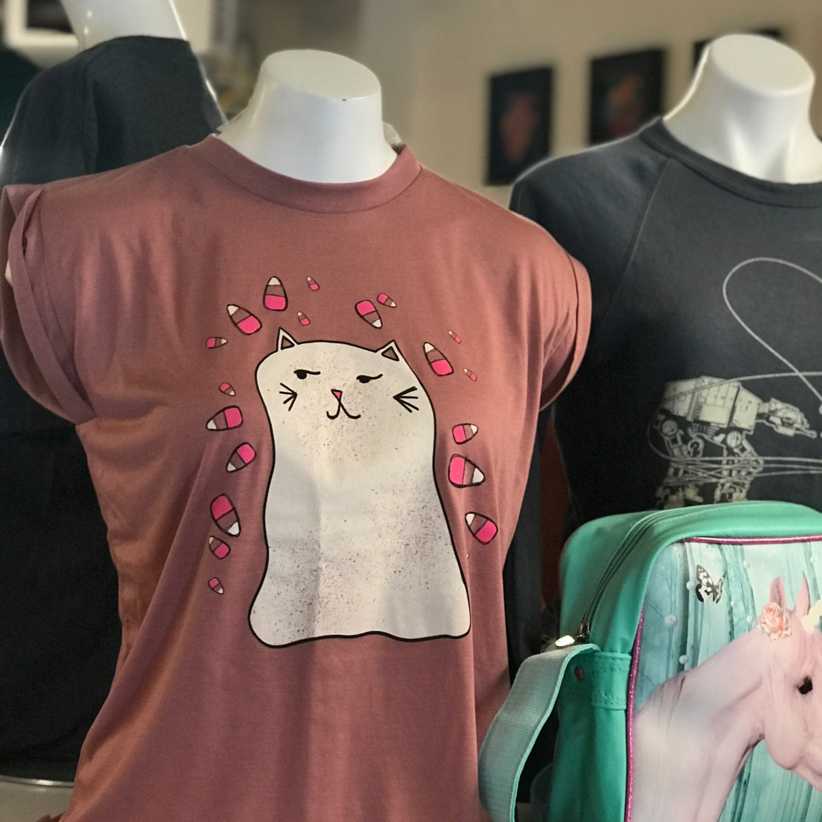 T-shirt with ghost cat design on a mannequin, next to a green bag with a pink unicorn design.