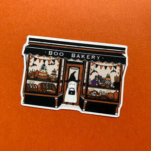 A die-cut sticker depicting a Halloween-themed bakery storefront with a ghost character in a witch hat standing in the doorway, placed on a textured orange background.