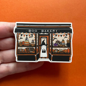 A hand holds a die-cut sticker or pin featuring a Halloween-themed bakery storefront with the words “BOO BAKERY” above windows displaying spooky treats, pumpkins, bats, and a ghost character in the center doorway, against an orange background.