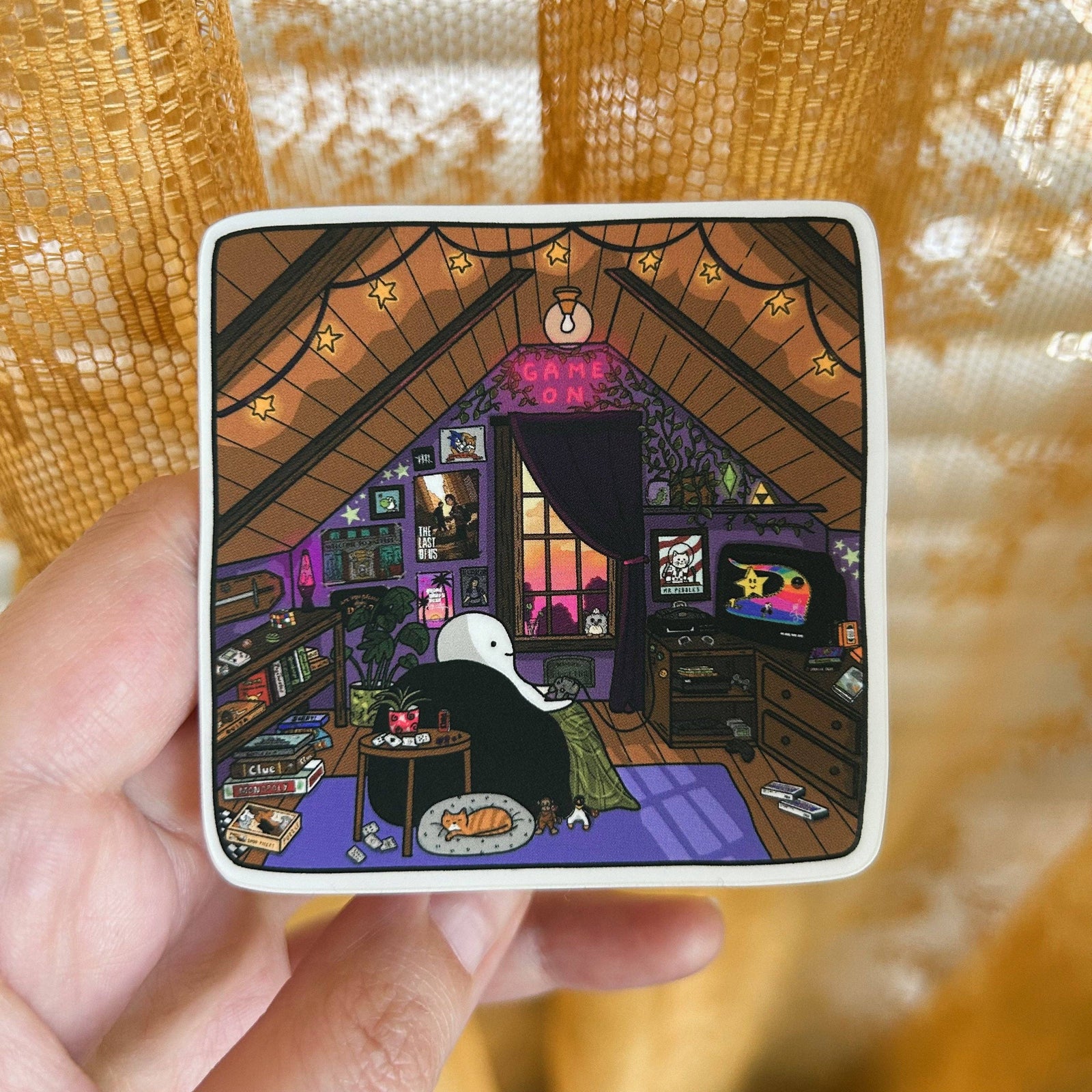 A hand holds a square white-bordered sticker featuring an illustrated cozy attic room scene with a black-and-white character sitting at a desk, surrounded by warm golden bokeh background and curtain-like texture.