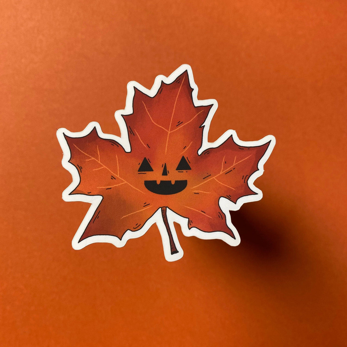Maple leaf with a jack-o'-lantern face on an orange background.