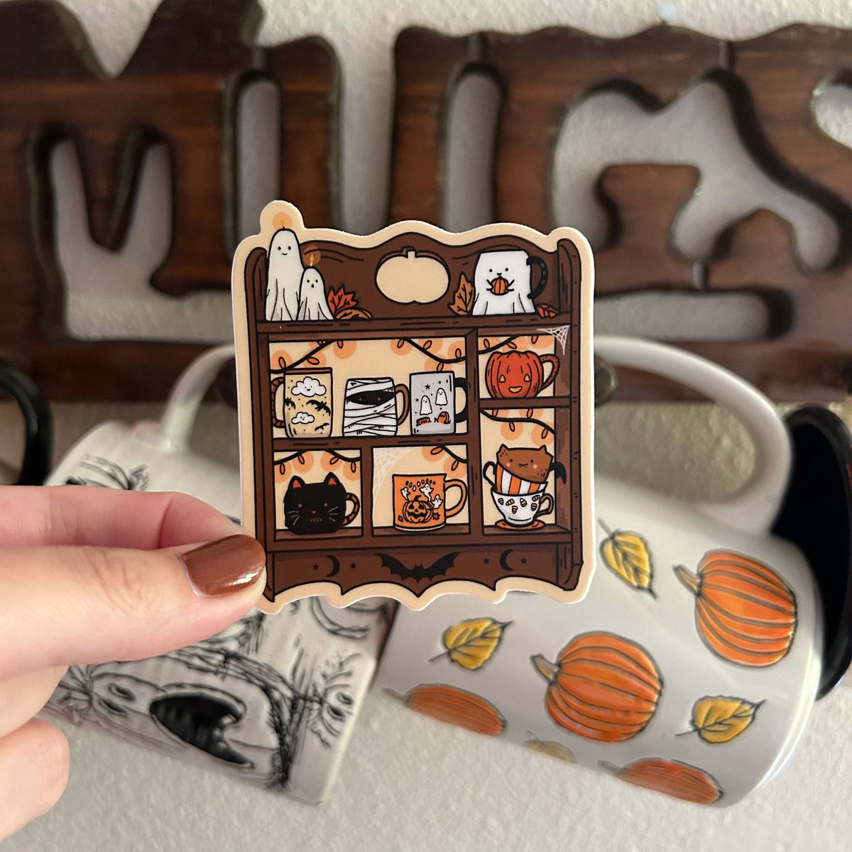 A hand with brown nail polish holds a die-cut sticker featuring a Halloween-themed wooden shelf with mugs, pumpkins, ghosts, and bats. In the background, a white ceramic mug with orange pumpkins and yellow leaves is visible, along with a dark wooden decorative sign.