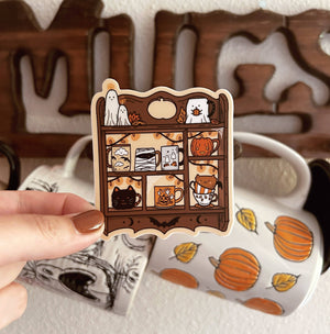 A hand holds a die-cut sticker featuring a whimsical shelf of Halloween-themed mugs and characters. In the background, a white ceramic mug with orange pumpkins and black cat illustrations is visible, along with a wooden decorative sign.