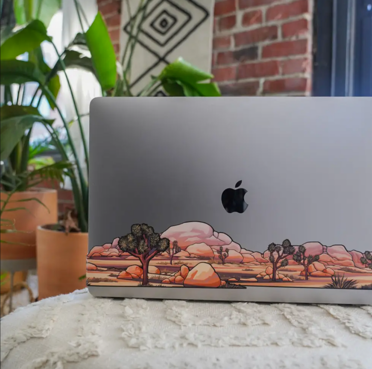 Laptop with a desert landscape decal on a surface with plants and a brick wall in the background