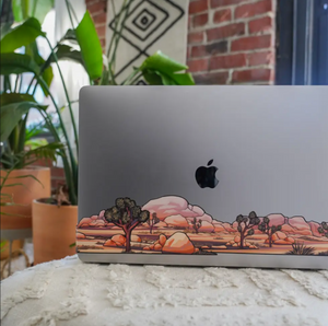 Laptop with a desert landscape decal on a surface with plants and a brick wall in the background
