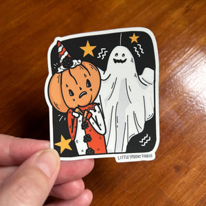 A hand holds a die-cut sticker featuring a cartoon pumpkin in an orange costume and a white ghost on a black background with gold stars, photographed over a wooden tabletop. The sticker reads 'LITTLE SPOOKY STUDIO'.