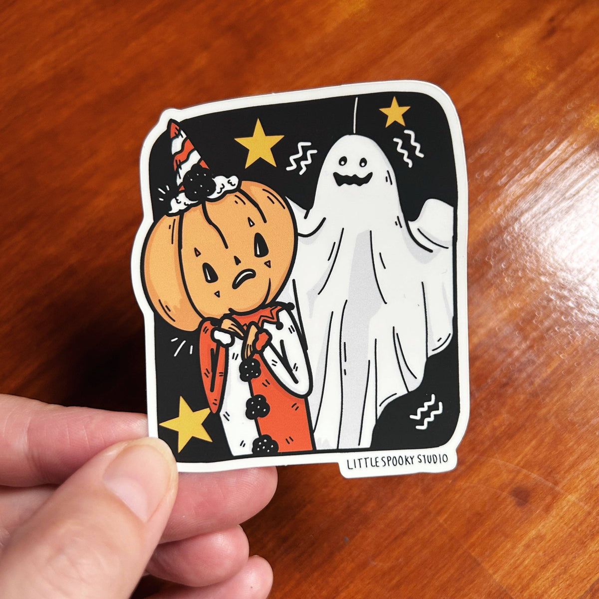 A hand holds a die-cut sticker featuring a Halloween illustration of a sad pumpkin and a ghost on a black background, with the text 'LITTLE SPOOKY STUDIO' at the bottom, set against a wooden table.