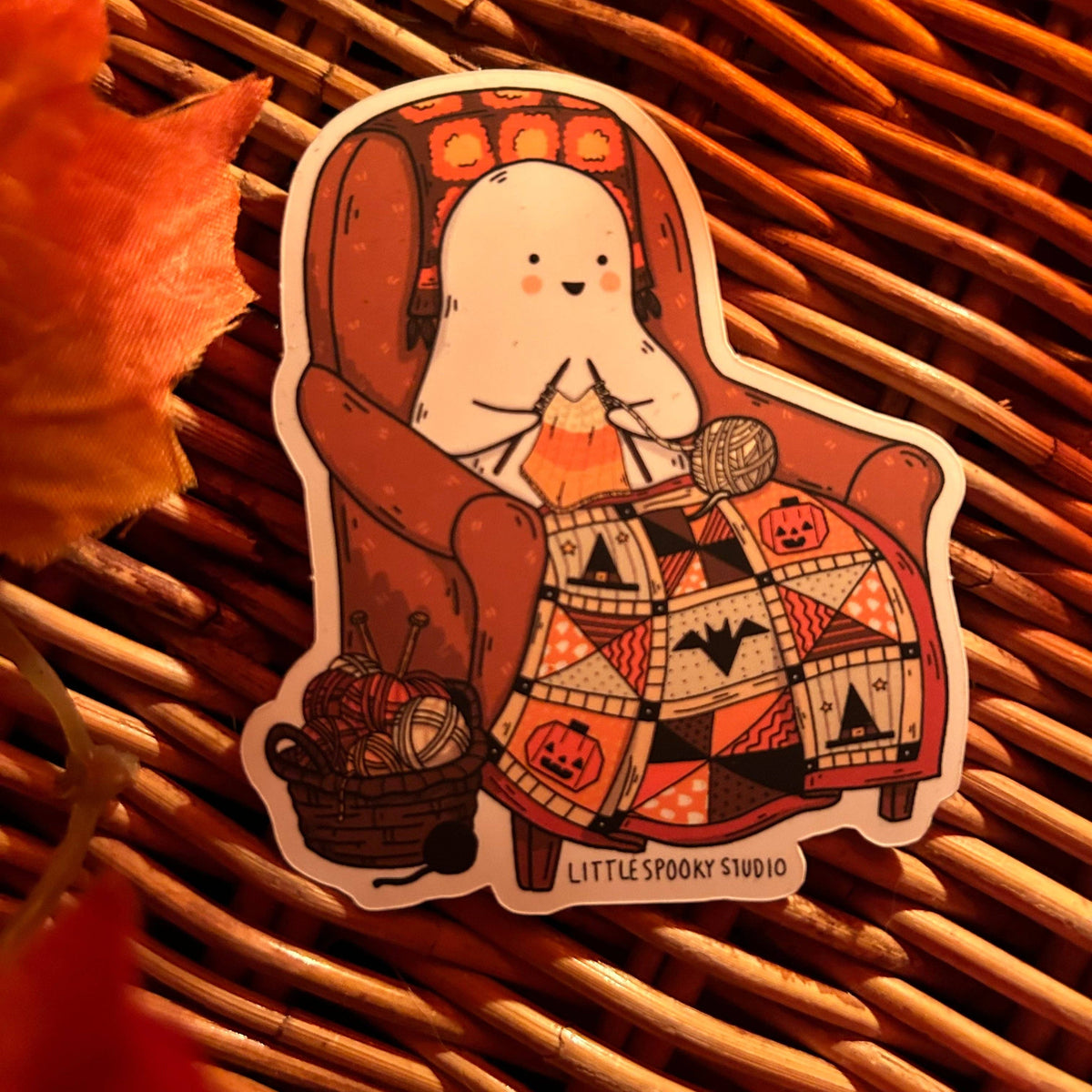 A die-cut sticker featuring a cute cartoon ghost sitting in a brown armchair knitting a patchwork blanket with Halloween motifs, placed on a brown wicker surface with a blurred orange leaf in the corner. The sticker reads 'LITTLE SPOOKY STUDIO'.
