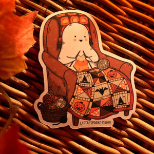 A die-cut sticker featuring a cute cartoon ghost sitting in a brown armchair knitting a patchwork blanket with Halloween motifs, placed on a brown wicker surface with a blurred orange leaf in the corner. The sticker reads 'LITTLE SPOOKY STUDIO'.