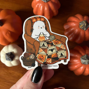 A die-cut sticker held by a hand with black nail polish, featuring a cute cartoon ghost sitting in a brown armchair knitting a patchwork blanket with pumpkins. The sticker is set against a background of small decorative pumpkins on a wooden surface.