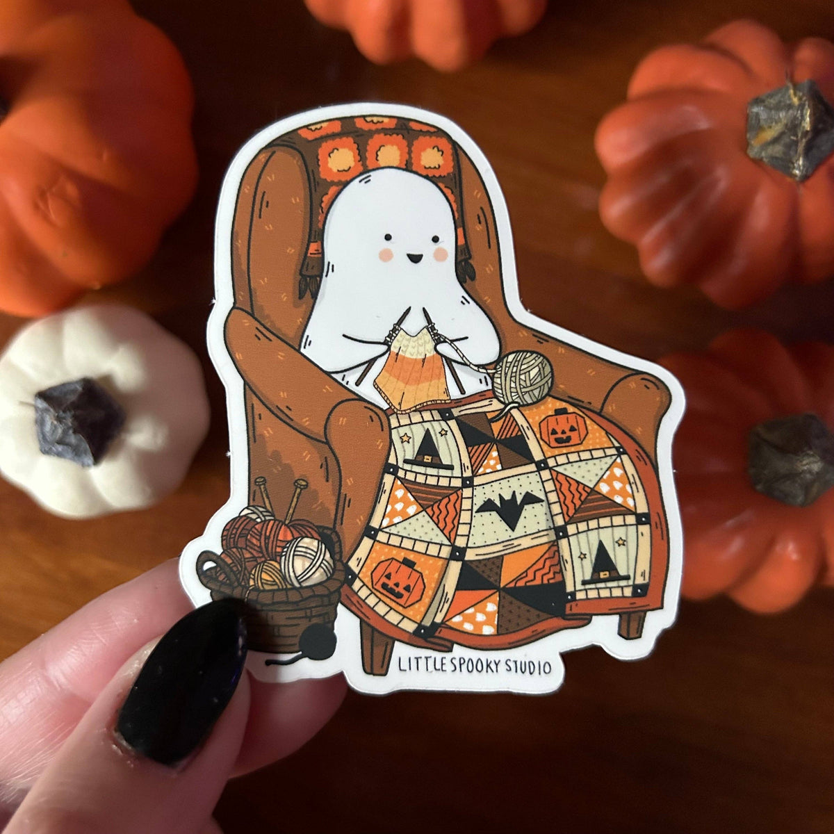 A die-cut sticker held by a hand with black nail polish, featuring a cute cartoon ghost knitting in a cozy armchair with a Halloween-themed patchwork blanket. The sticker is set against a warm, blurred background of orange pumpkins.