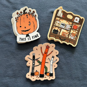 Three Halloween-themed die-cut stickers displayed on a textured slate blue fabric background. The stickers feature whimsical illustrations: a smiling orange pumpkin with mushrooms growing out of its head and the text 'THIS IS FINE', a haunted house shelf scene with cute ghostly characters, and three stick-like figures (black, orange, and white) with arms raised under falling leaves.