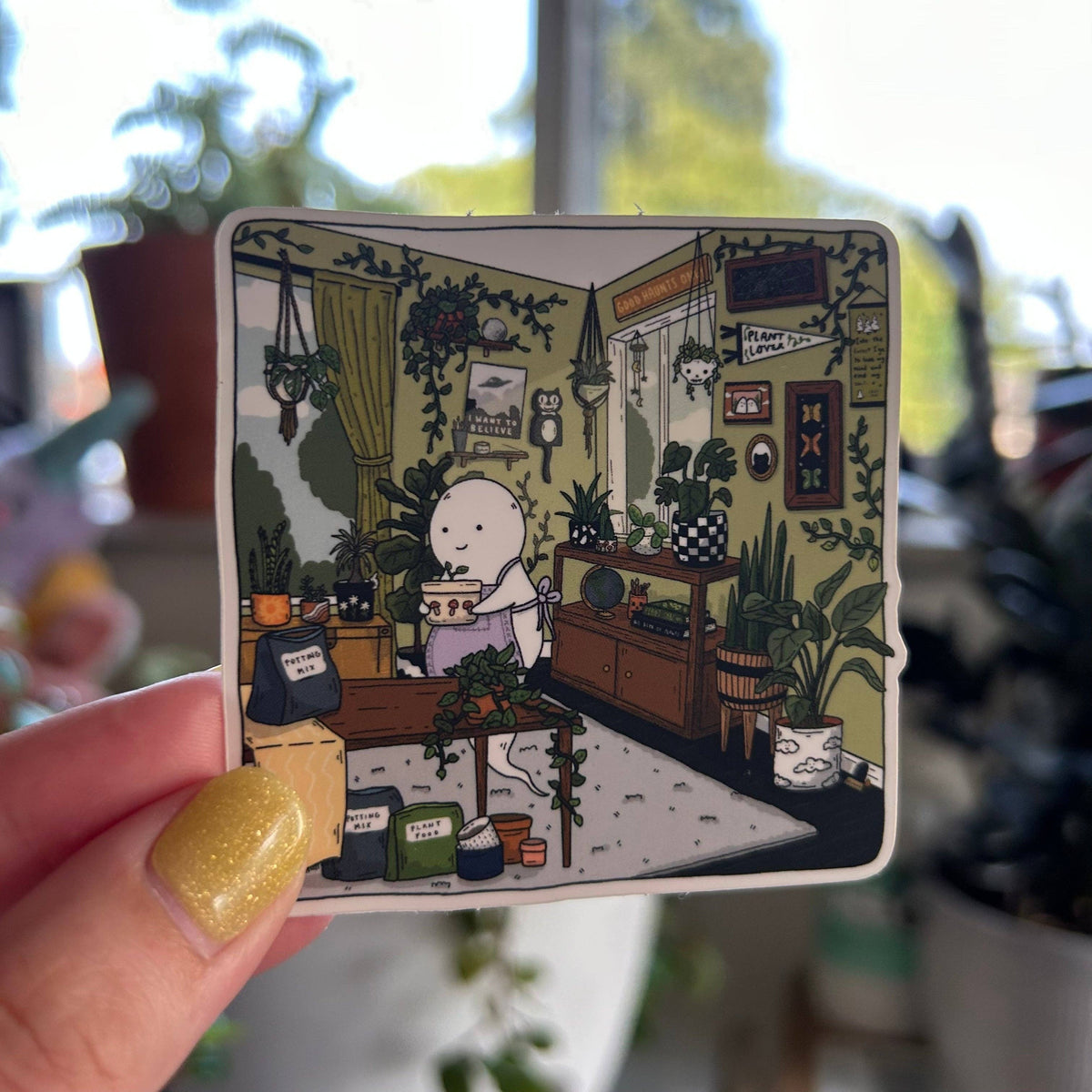 A hand with glittery yellow nails holds a square sticker featuring a whimsical illustration of a small white character in a cozy plant-filled room. The background is a blurred indoor setting with potted plants and bright natural light.