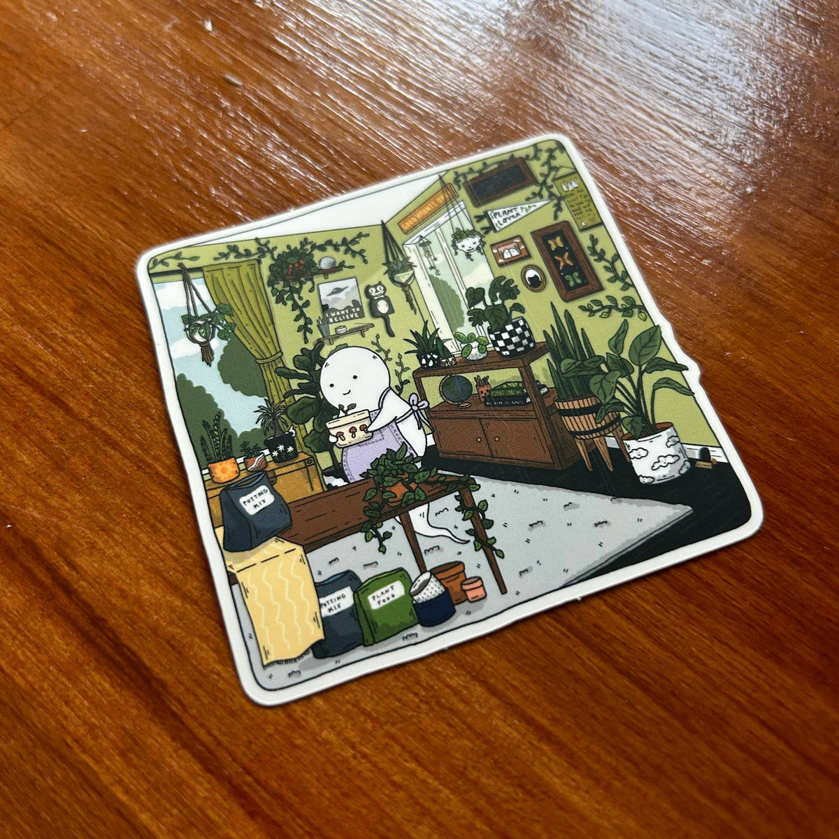 A square die-cut sticker with rounded corners featuring a detailed cartoon illustration of a white-haired character in a cluttered room with plants and coffee mugs, placed on a brown wooden table.