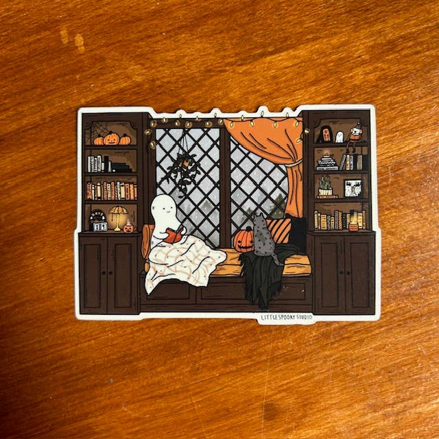 A rectangular die-cut sticker featuring a cozy Halloween-themed illustration of a ghost and cat on a window seat, placed on a wooden tabletop.