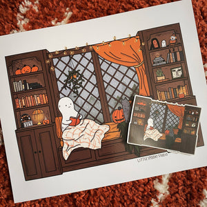 A printed art card or poster featuring a cozy Halloween illustration of a ghost reading in a window seat, surrounded by pumpkins, books, and orange curtains, placed on a textured red-orange rug with a smaller photo printed on top.