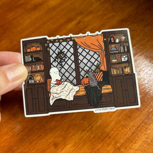 A hand holds a die-cut sticker featuring a cozy Halloween scene with a ghost in a bed, a cat, bookshelves, and pumpkins. The sticker has a white border and small text at the bottom reading 'LITTLE SPOOKY STICKER'.