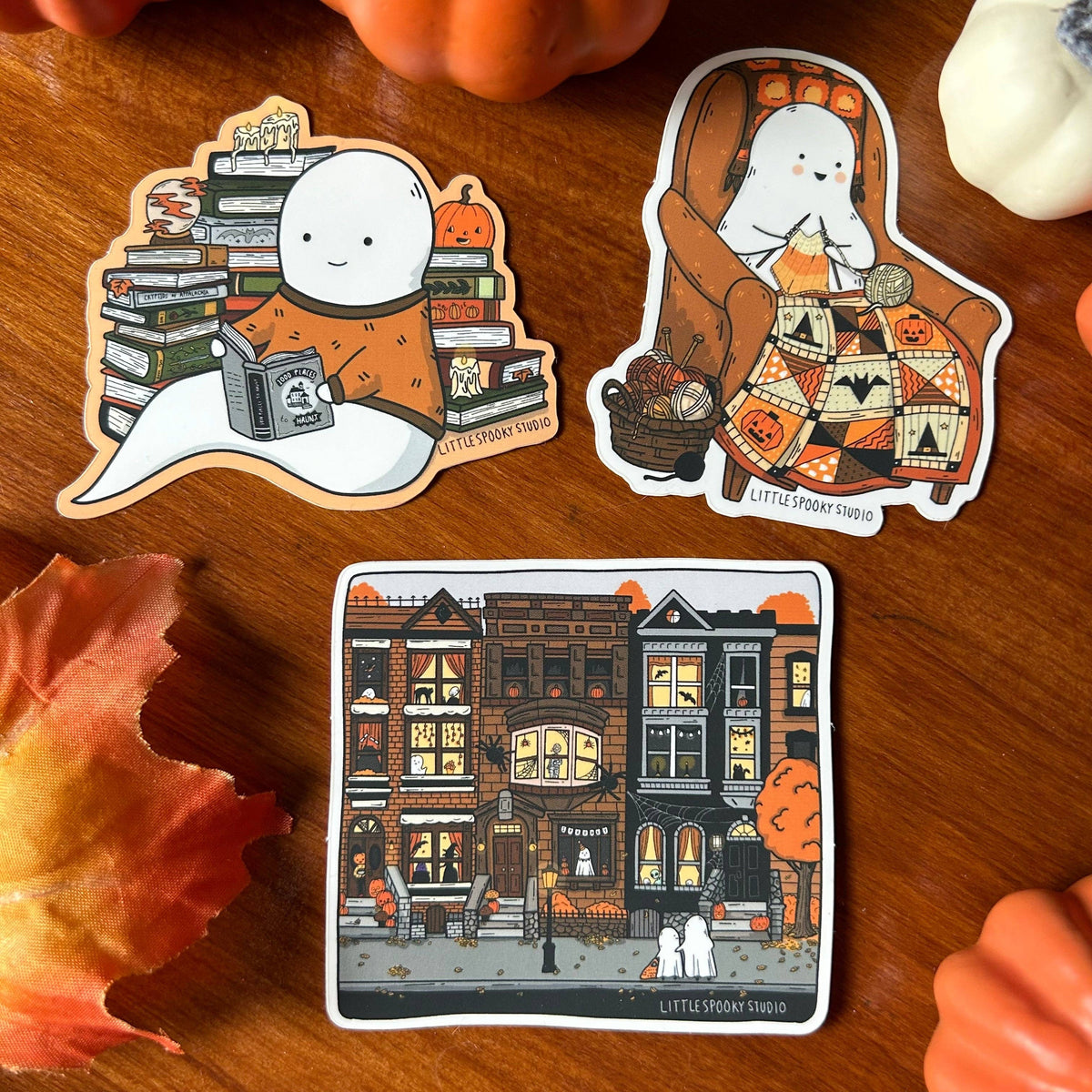 Three die-cut stickers featuring cartoon ghosts and a cozy autumn street scene, arranged on a wooden surface with fall props. The stickers include a ghost reading under stacked books, a ghost sitting in an armchair with a patterned blanket, and a square illustration of haunted houses along the street.