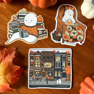Three die-cut stickers featuring cartoon ghosts and a cozy autumn street scene, arranged on a wooden surface with fall props. The stickers include a ghost reading under stacked books, a ghost sitting in an armchair with a patterned blanket, and a square illustration of haunted houses along the street.