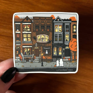 A hand with black nail polish holds a square white sticker featuring a stylized illustration of spooky townhouses, pumpkins, cobwebs, and two ghosts. The sticker has the text 'LITTLESPOOKY STUDIO' at the bottom and is photographed on a wooden surface.