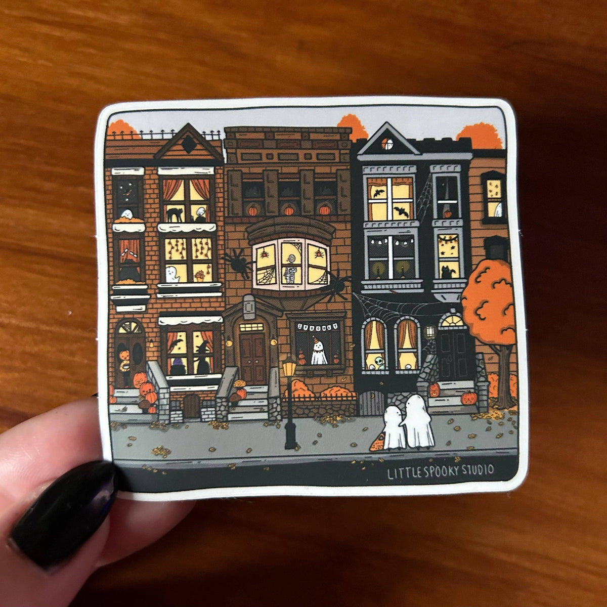A square sticker featuring a whimsical illustration of three haunted Victorian townhouses with glowing windows, pumpkins, and two white ghosts, held by a hand with black fingernails against a wooden background. The bottom right corner of the sticker reads 'LITTLE SPOOKY STUDIO'.