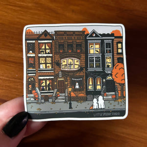 A square sticker featuring a whimsical illustration of three haunted Victorian townhouses with glowing windows, pumpkins, and two white ghosts, held by a hand with black fingernails against a wooden background. The bottom right corner of the sticker reads 'LITTLE SPOOKY STUDIO'.