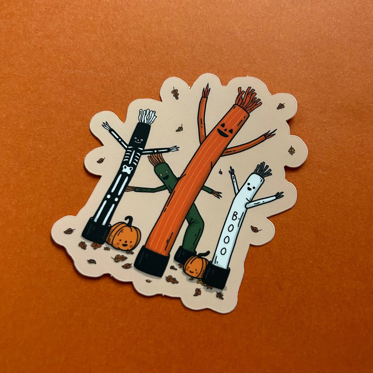 A die-cut sticker featuring three cartoon Halloween figures and pumpkins on an orange background.