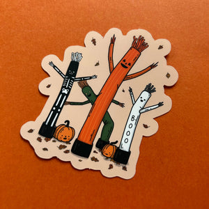 A die-cut sticker featuring three cartoon inflatable bouncy figures in black and white, orange, and green, along with two jack-o'-lanterns, shown on an orange textured background.