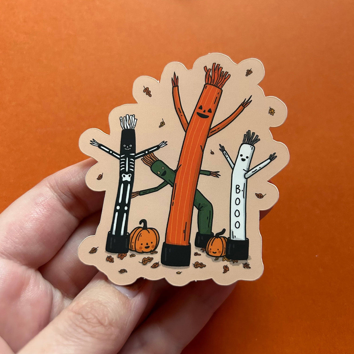 A hand holds a scalloped die-cut sticker featuring three Halloween-themed stick figures: a skeleton, a tall orange tree-like figure, and a white figure with 'BOO' written on it, with small pumpkins at the bottom. The background is a solid orange surface.