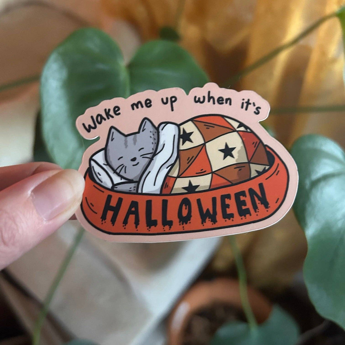 Sticker with a cat design and text 'Wake me up when it's Halloween' held by a hand with a plant in the background.