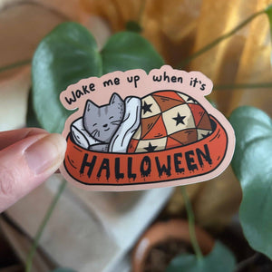 Sticker with a cat design and text 'Wake me up when it's Halloween' held by a hand with a plant in the background.