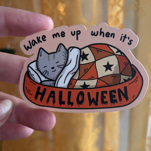 Sticker with a cat and text 'Wake me up when it's Halloween' held by a hand.