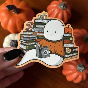 A die-cut sticker featuring a cartoon ghost reading books, held by a hand with black nail polish against a background of real pumpkins. The sticker has 'LITTLESPOOKY STUDIO' printed on the bottom right.