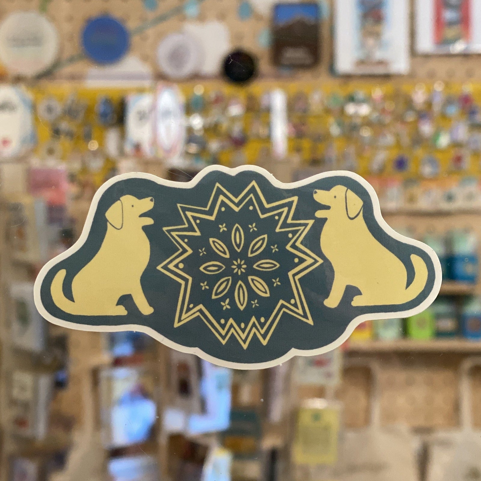 A folk art style sticker with two lab dogs sitting facing each other. A folk art design is between then. The sticker is blue and off-white.
