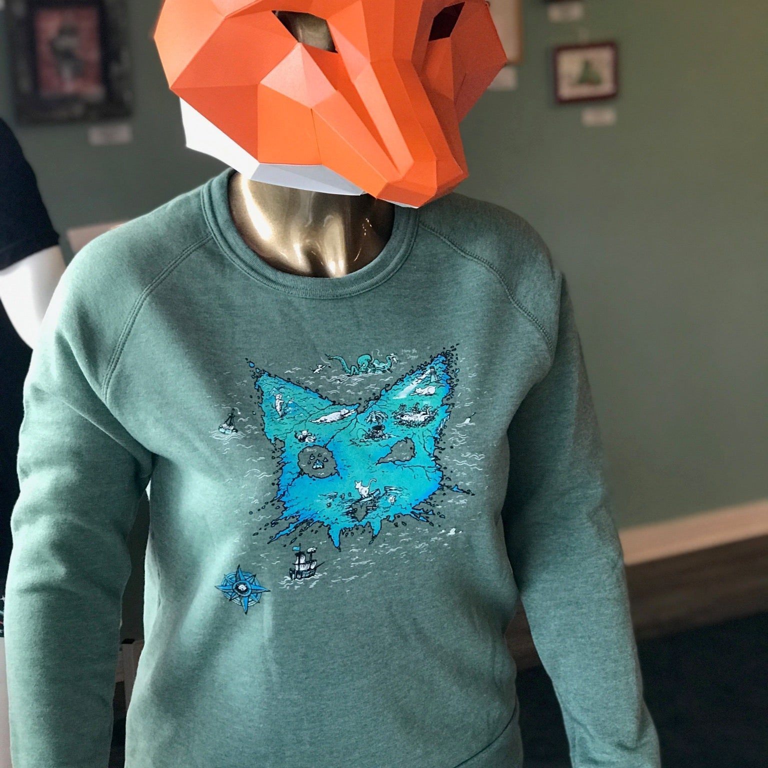 A person in a fox mask is wearing a green sweatshirt with a cat head shaped treasure map on the front. 