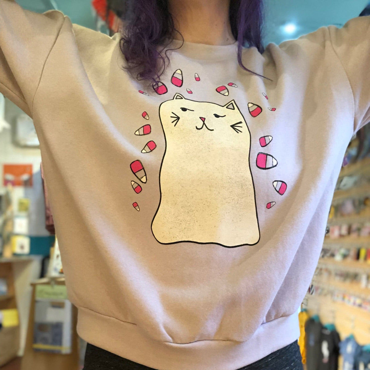 Person wearing a blush pink sweatshirt with a cartoon ghost cat design in a store setting. 