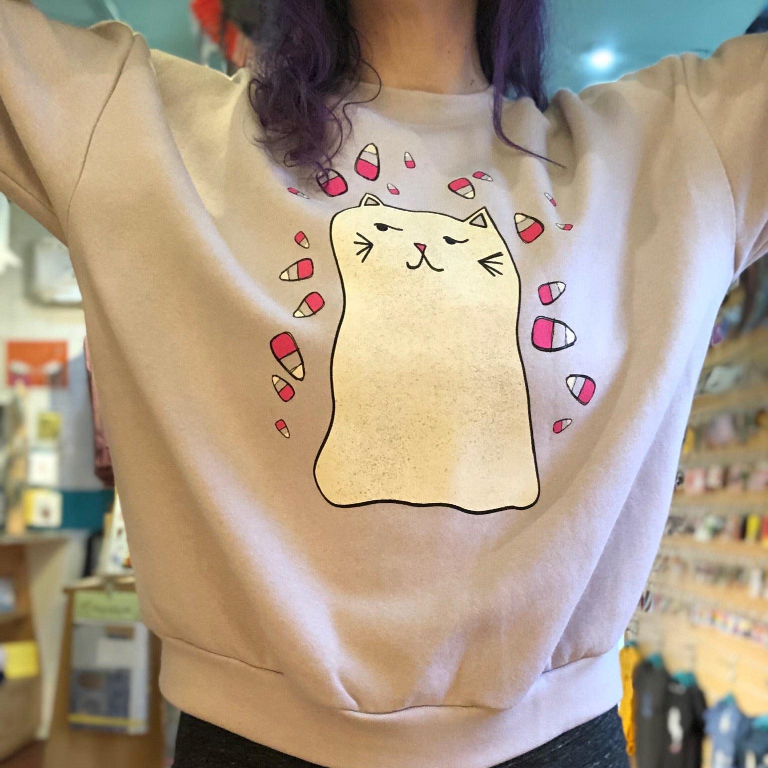 Person wearing a blush pink sweatshirt with a cartoon ghost cat design in a store setting. 