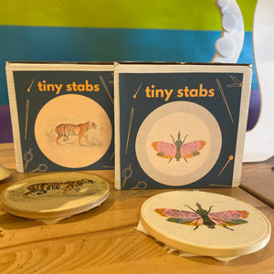 Two 'tiny stabs' embroidery kits with illustrations of a tiger and a moth on a wooden surface.
