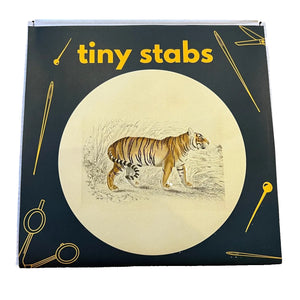 A square product box that says "Tiny Stabs" on the front. The box features a dark forest green background with a large cream-colored circular illustration in the center showing a tiger, surrounded by yellow line drawings of sewing tools.