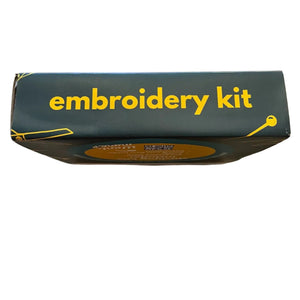 A dark forest freen cardboard box labeled 'embroidery kit' in yellow text, photographed from above on a worn wooden tabletop.